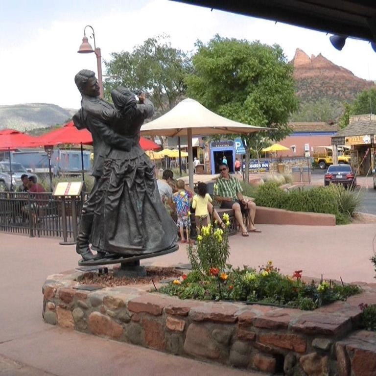 Eine Statue von drei Figuren steht in einem öffentlichen Platz, umgeben von Geschäften und mit bergigem Panorama im Hintergrund.