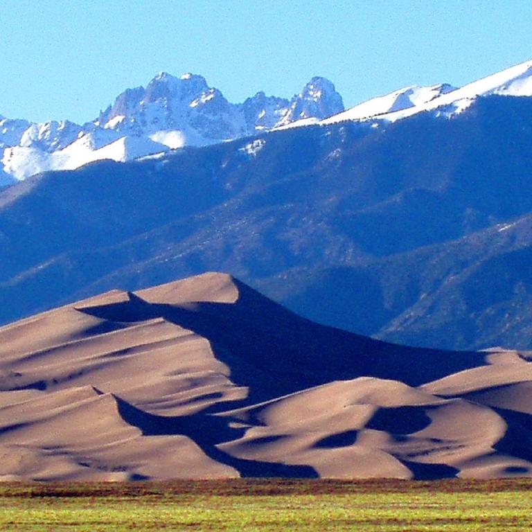 Eine Sanddünenlandschaft vor einem beeindruckenden Bergpanorama mit schneebedeckten Gipfeln.