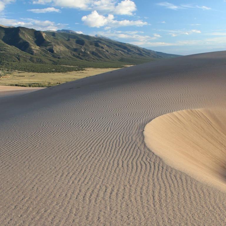 Großer Sanddünenkopf mit feinen Wellenstrukturen und dahinter sanfte grüne Bergkette unter blauem Himmel.
