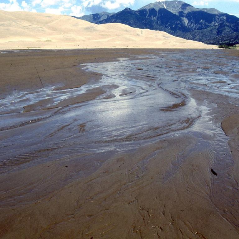 Ein flaches Gewässer mit braunem Schlammboden und Sedimentfahnen vor einer Sanddüne und Bergen.