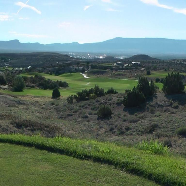 Panoramaaufnahme eines Golfplatzes mit trockenen, bewachsenen Hügeln und majestätischen Bergen im Hintergrund.