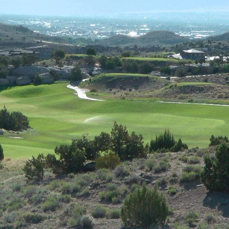 Panoramaaufnahme eines weiten Golfplatzareals, das von trockener Buschvegetation eingerahmt wird und in eine Siedlung mit hügeligem Hintergrund überge
