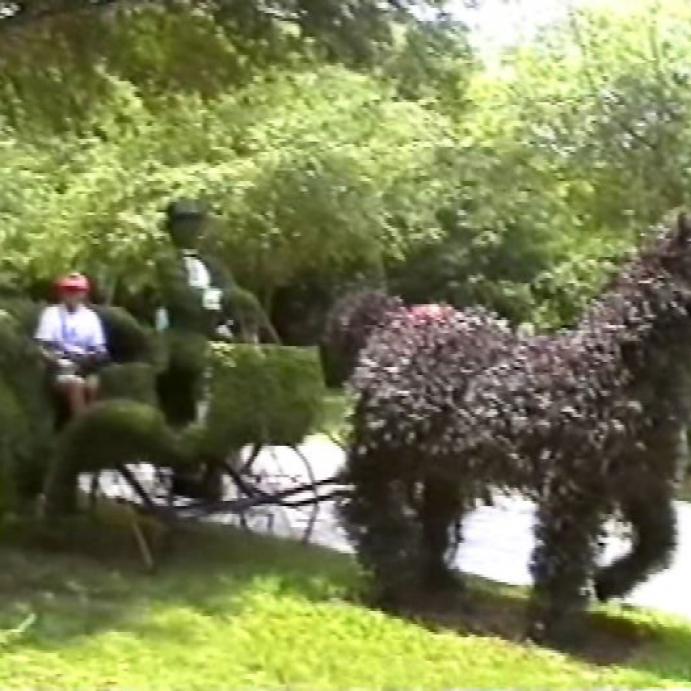 Ein buschartiger, dunkelgrüner Charakter marschiert auf einem Weg durch einen parkähnlichen Bereich, während links ein Kinderwagen zu erkennen ist.