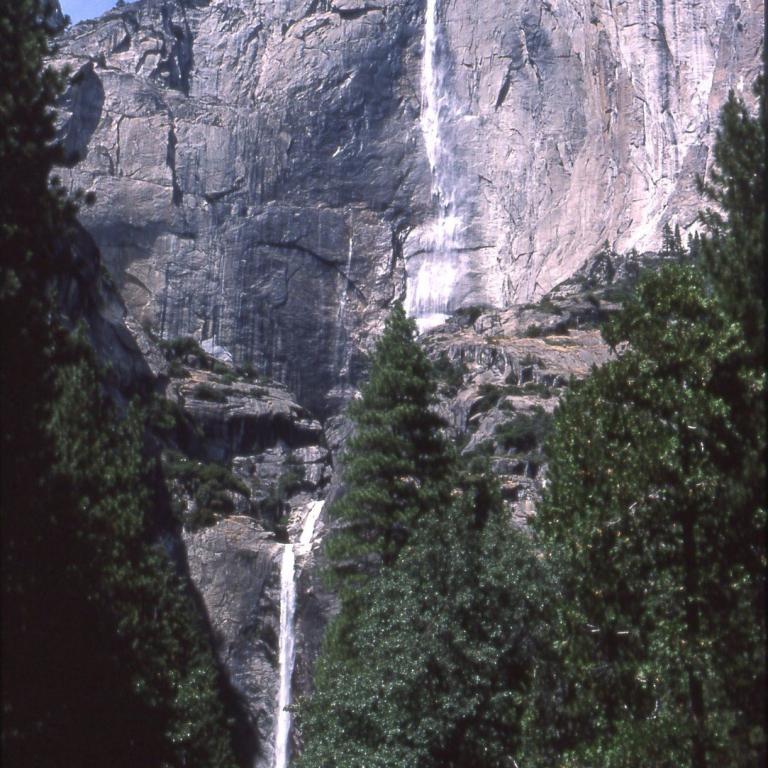 Ein großer Wasserfall stürzt von einer massiven, grauen Felswand herab und wird von dunklen Nadelbäumen im Vordergrund eingerahmt.