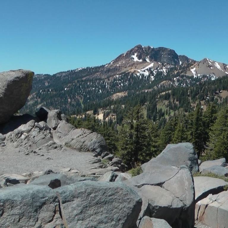 Panorama einer Berglandschaft mit großen Felsen im Vordergrund, dichtem Nadelwald und majestätischen Gipfeln unter blauem Himmel.