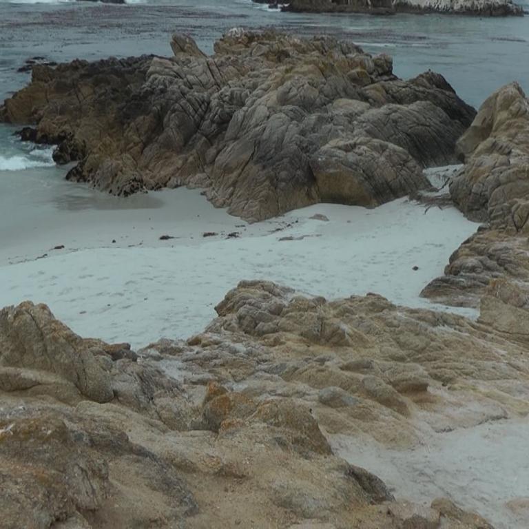 Steile, felsige Küste mit breitem, sandigem Strand im Hintergrund.