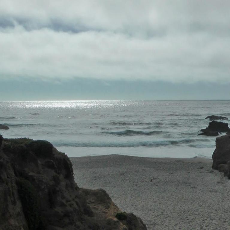 Küstenlandschaft mit steilen Klippen, einem Sandstrand und dem Meer unter bewölktem Himmel.