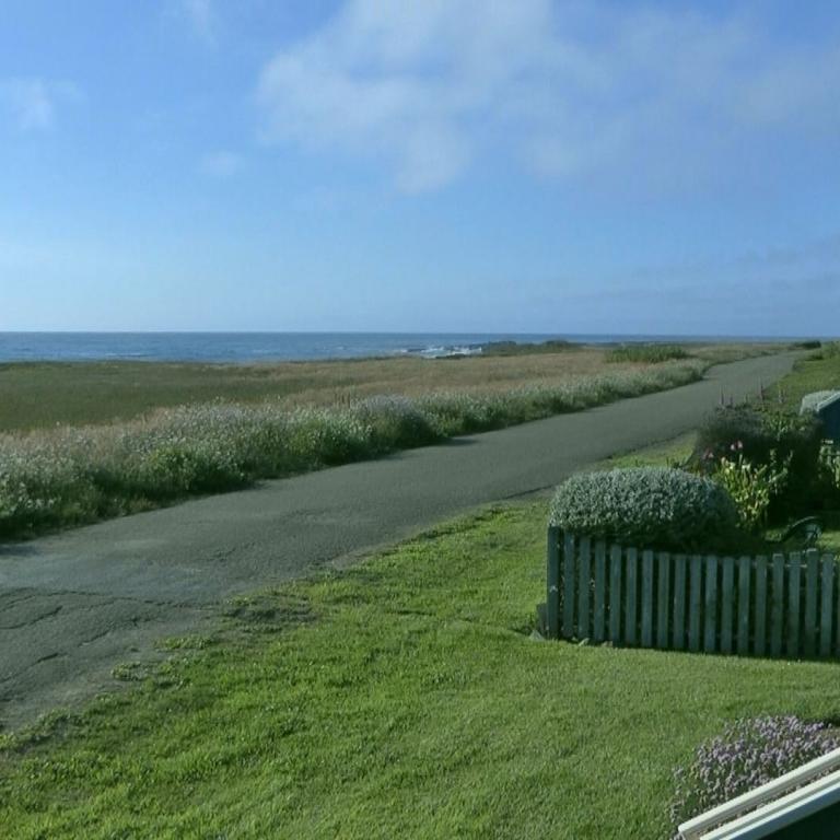 Ein gepflasterter Küstenweg führt durch Dünenvegetation mit Blick auf das Meer und einem modernen Gebäude rechts.