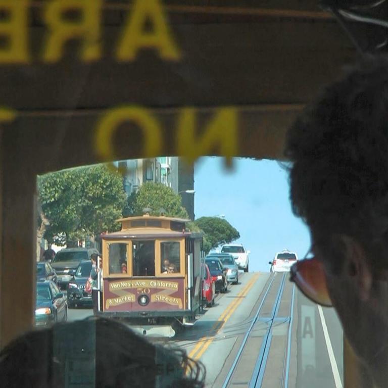 Ansicht von einem Auto, die eine historische Straßenbahn und andere Fahrzeuge in einer belebten städtischen Umgebung zeigen, wobei ein unscharfer Betr