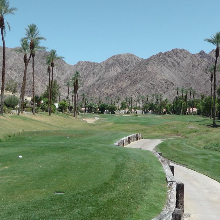 Panoramablick auf einen Golfplatz mit einem Pfad im Vordergrund, Palmen und trockenen Bergketten im Hintergrund.