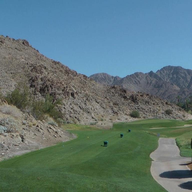 Ein Golfplatz mit perfekt gepflegter Rasenfläche und einem Weg im Vordergrund, dahinter steigt eine trockene Bergkette an.