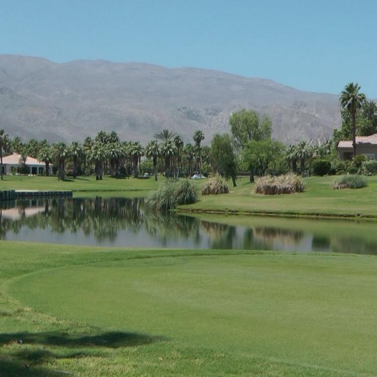 Ein Golfplatz mit Teich und Palmen im Vordergrund, dahinter Villen und einer ausgedehnten Bergkette im Hintergrund.