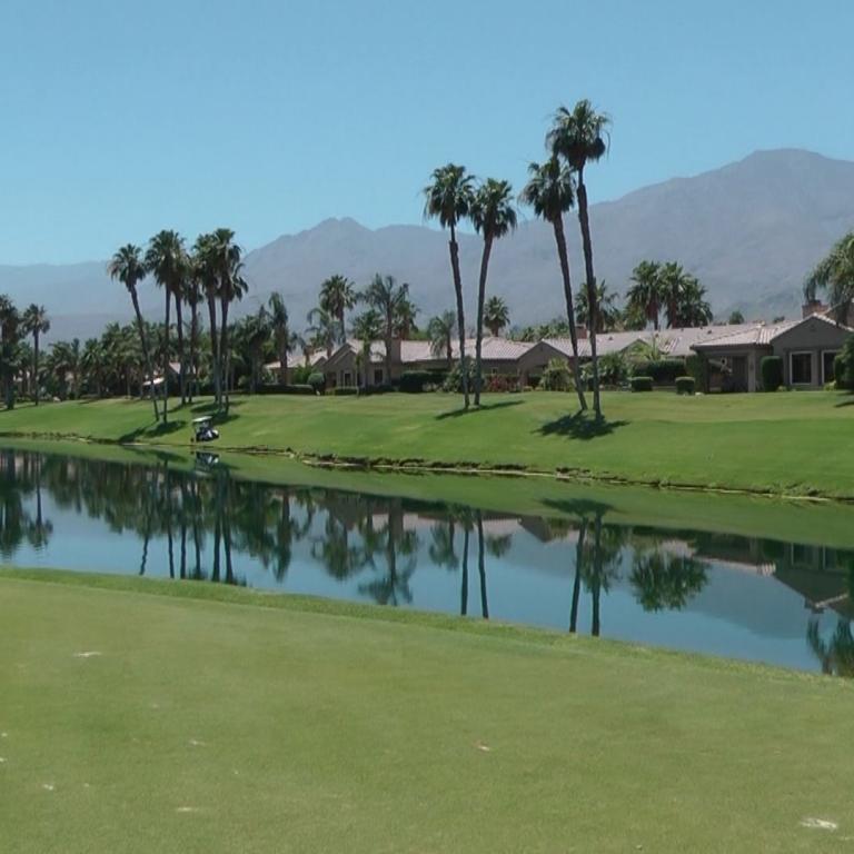 Blick auf einen grünen Golfplatz mit einem reflektierenden Wasserteich, umrahmt von Palmen und Bergen.