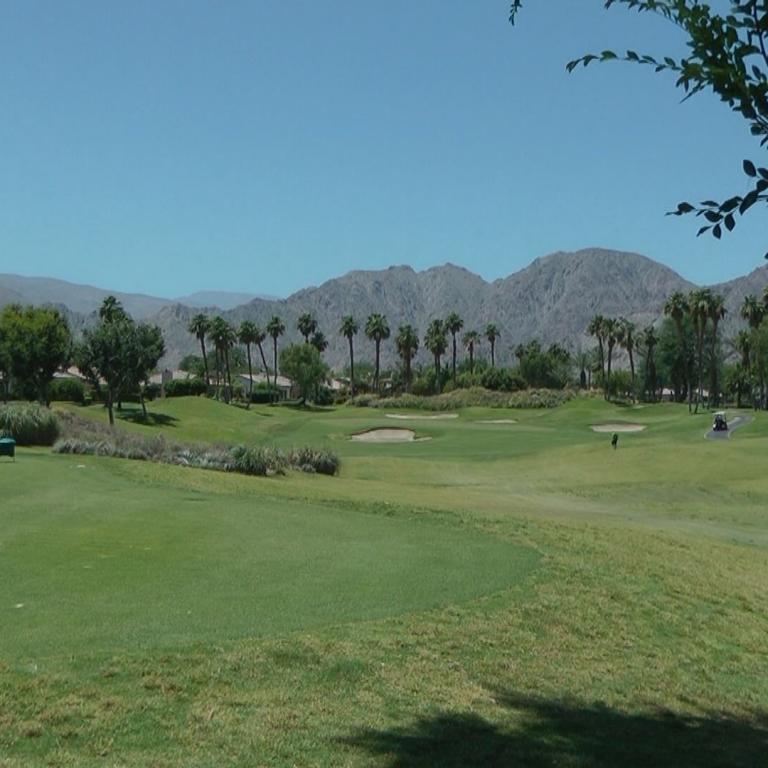 Panoramaansicht eines Golfplatzes mit Bergkulisse und Bäumen im Vordergrund.