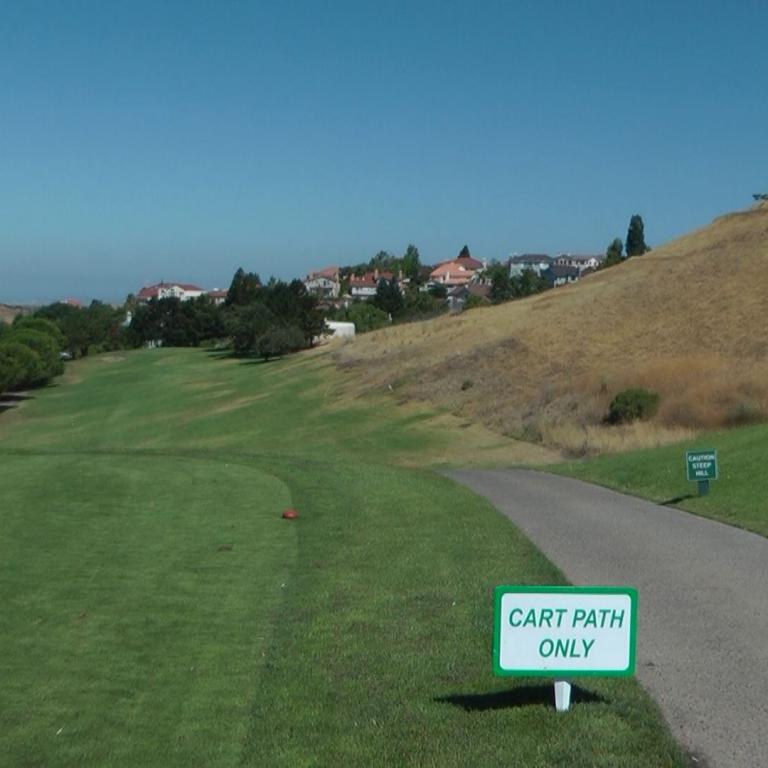 Panoramaansicht eines Golfplatzes mit grünem Vordergrund, einem befestigten Wegen und einem steil abfallenden, trockenen Hang.