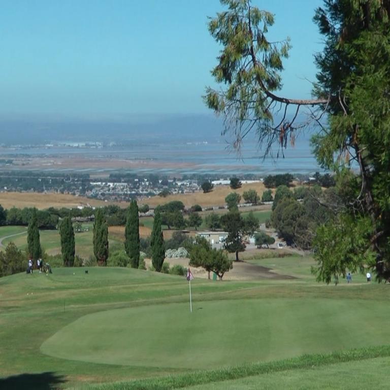 Panoramablick auf eine weite städtische Landschaft von einem Golfplatz aus, umrahmt von Bäumen.