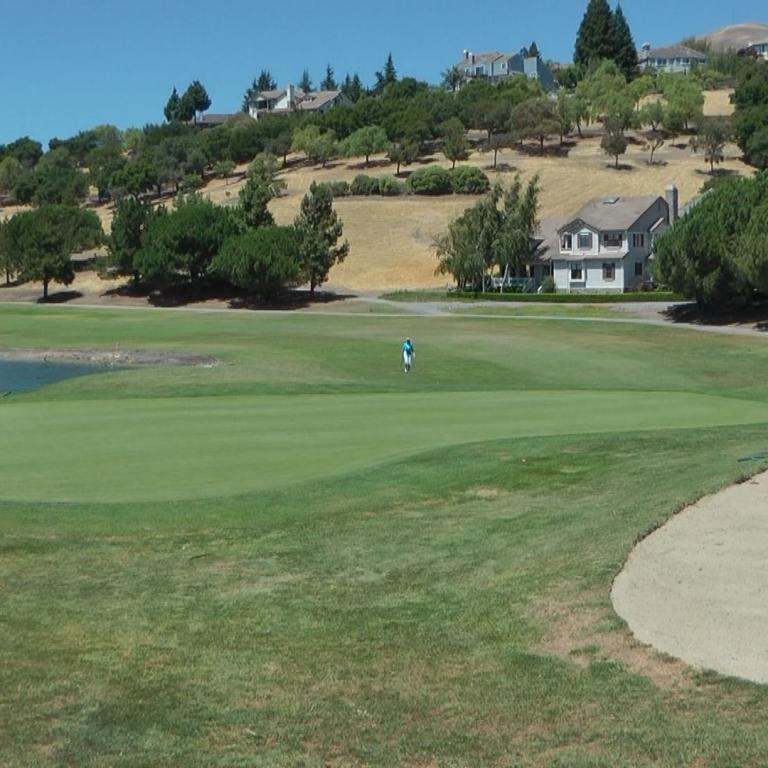 Eine weite Golfplatzszene mit einem Wasserloch im Vordergrund, einem Sandbunker rechts und Wohnhäusern auf einem bewaldeten Hang im Hintergrund.