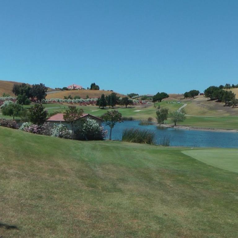 Panoramablick auf einen grünen Golfplatz mit Wasserteich vor einer Kulisse von hügeliger, baumbesetzter Landschaft.
