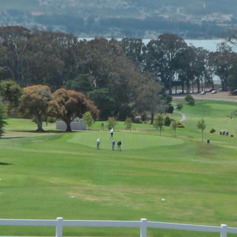 Ein weites Panorama eines Golfplatzes mit Spielern, dahinter ein See und bewaldete Hügel.
