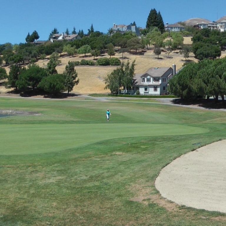 Weitwinkelaufnahme eines Golfplatzes mit einem Haus am Hang und Bäumen im Hintergrund.