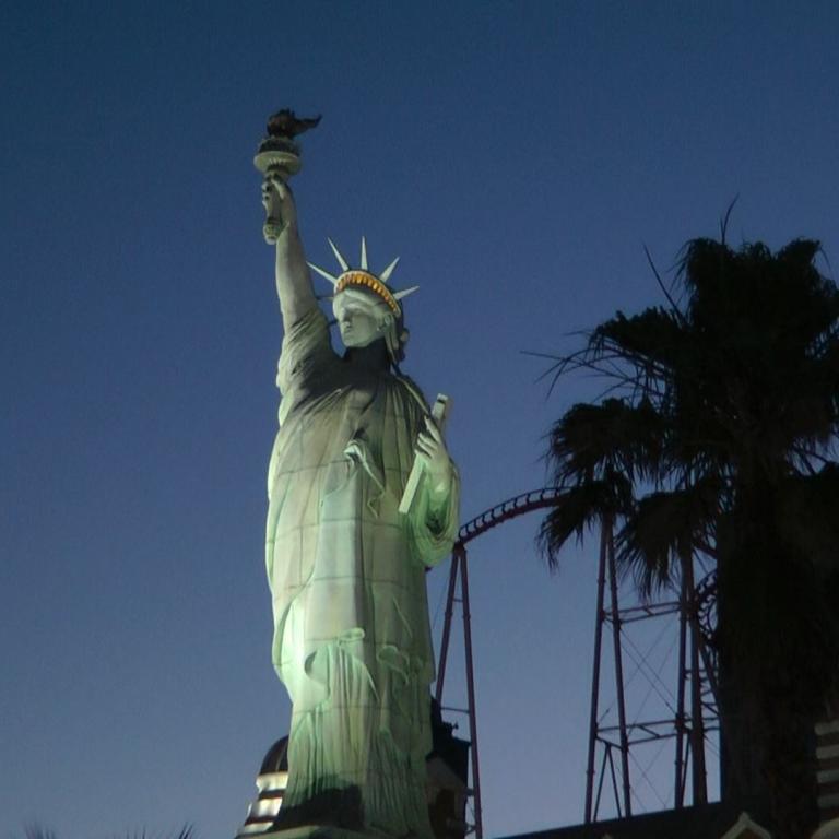 Die Freiheitsstatue steht vor einem tiefblauen Himmel, mit der Skyline New Yorks und Palmen im Hintergrund.