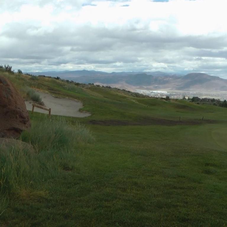 Panoramablick auf einen grünen Golfplatz mit einem Erdwall und Blick auf die Hügel unter einem bewölkten Himmel.