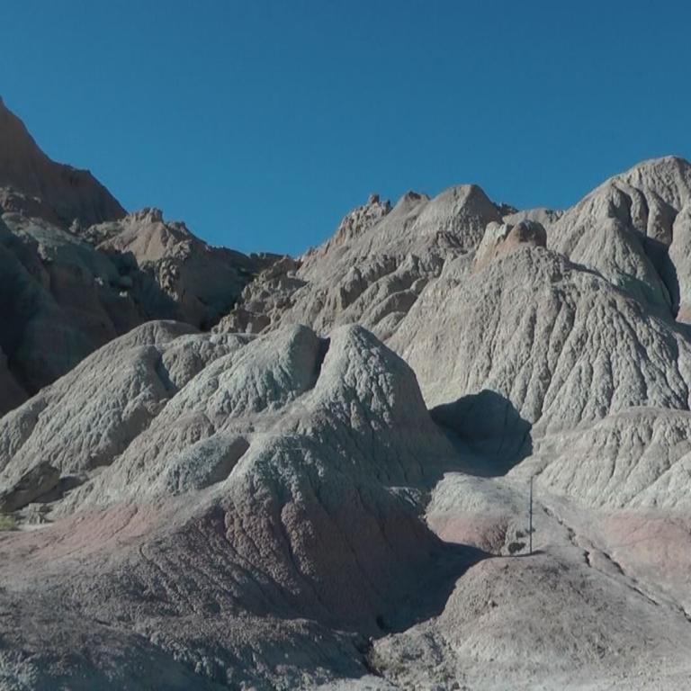 Eine aride Hügellandschaft mit erodierten Felsformationen in Beige-, Rosa- und Brauntönen unter einem klaren blauen Himmel.