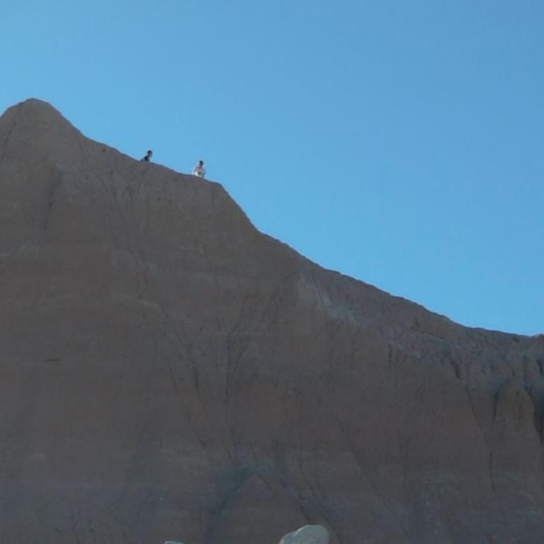 Eine große, braune Bergformation ragt unter einem klaren blauen Himmel empor mit zwei kleinen menschlichen Figuren auf dem Gipfel.