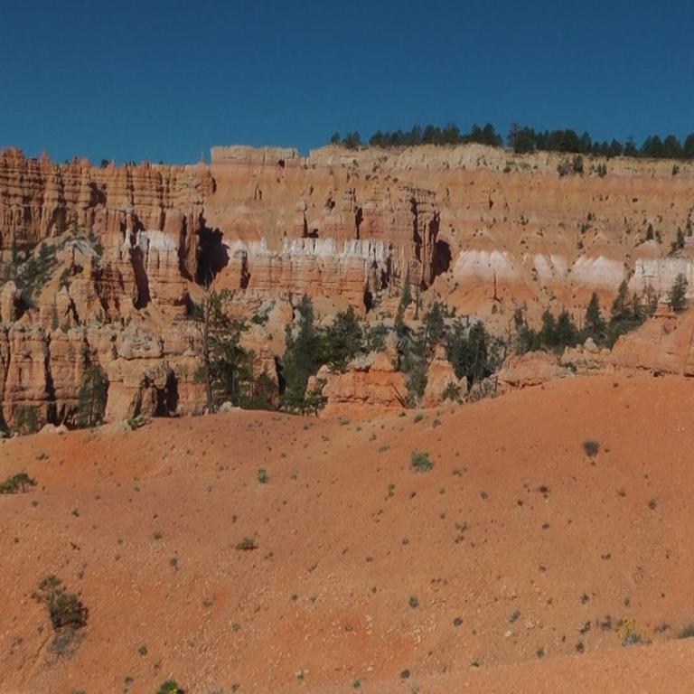 Eine weite aride Landschaft mit geschichteten roten Felskliffs und Vegetation unter blauem Himmel.