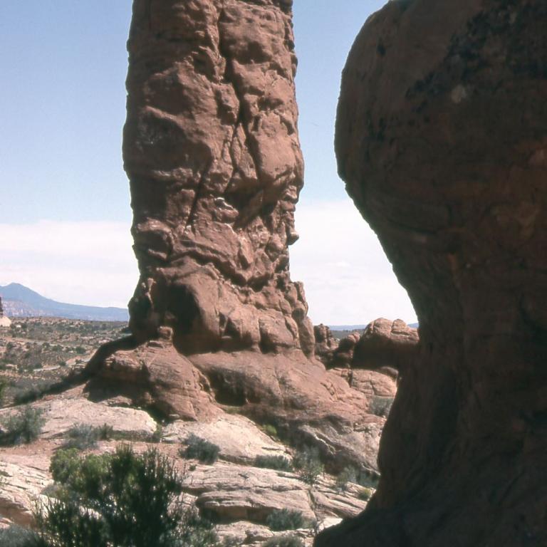 Ein hoher, markanter Felsmonolith steht in einer weiten Wüstenlandschaft, rechts davon ist eine weitere große Felsformation sichtbar.